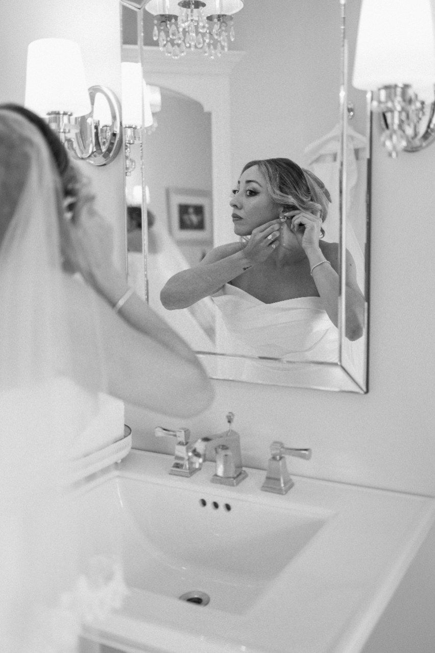 Black and white photo of someone putting on earrings while looking in a bathroom mirror with an elegant chandelier above.
