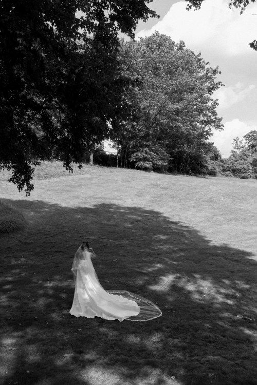 A long white wedding veil trails across a grassy hill in black and white with trees in the background.