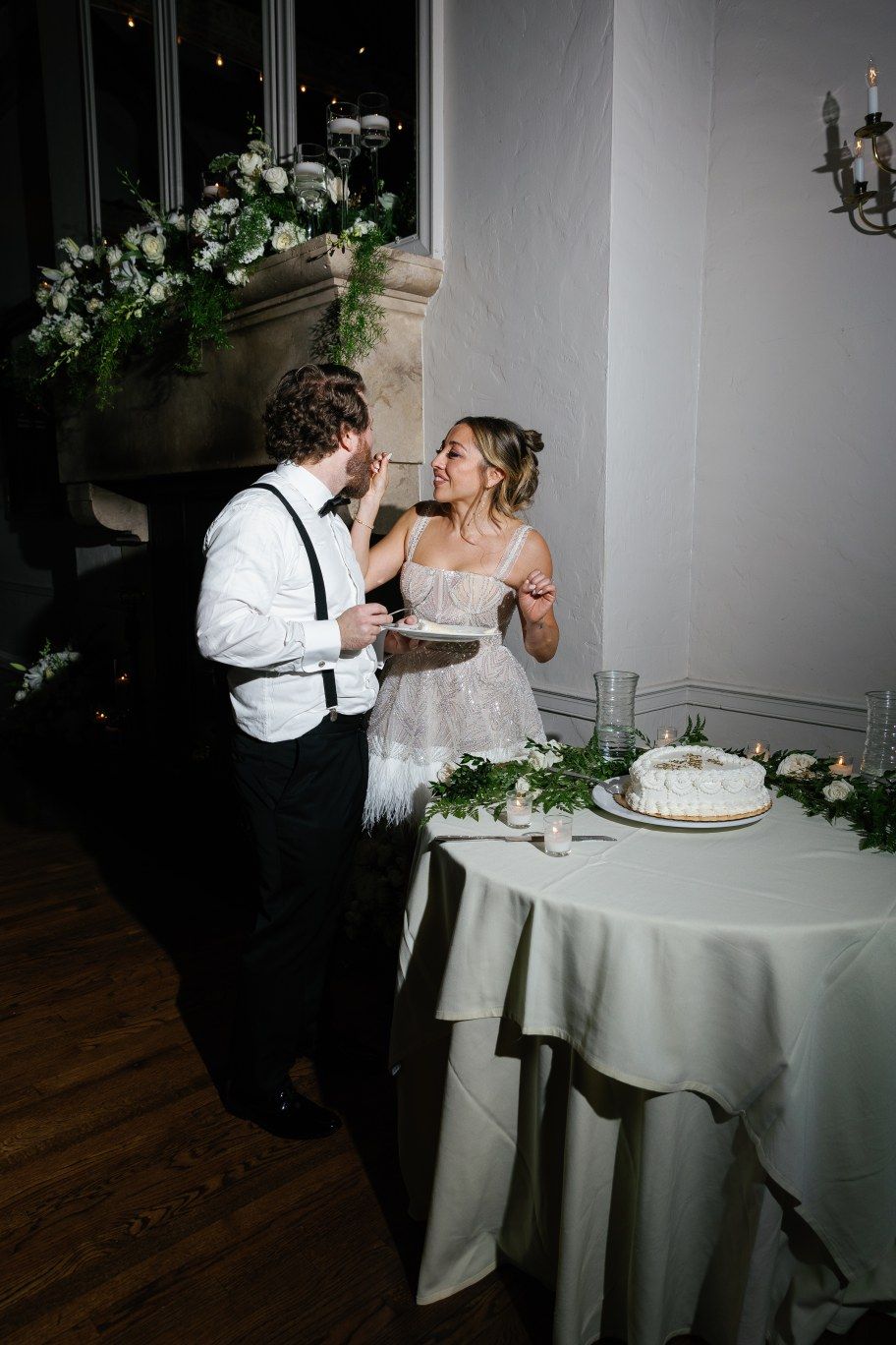 A bride and groom share cake-feeding moments at their wedding reception around a white tablecloth-covered table.