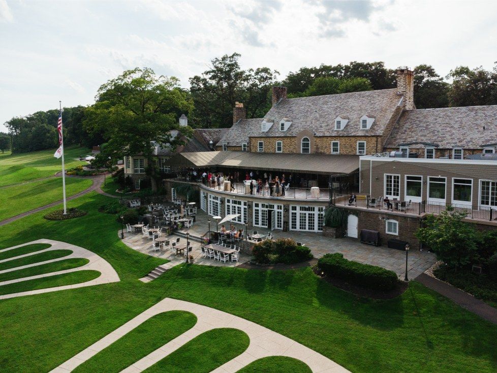 Aerial view shows a grand mansion estate with winding walkways and manicured lawns surrounded by lush landscaping.