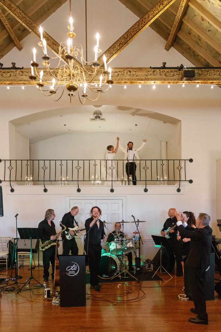Wedding band performs in elegant venue with chandelier and balcony while guests celebrate above on the mezzanine level.