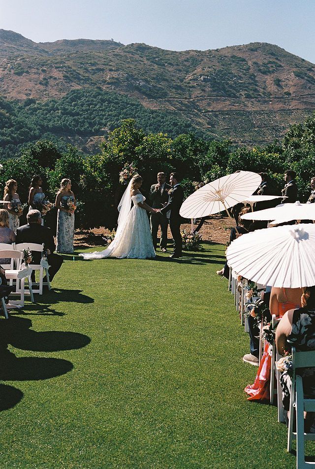 Outdoor wedding ceremony on a grassy lawn with white parasols and mountain backdrop.