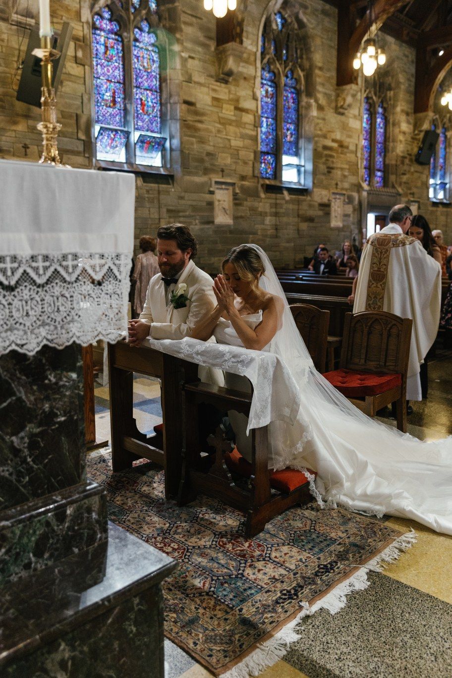 A couple in wedding attire prays at an ornate church altar with stained glass windows and stone walls in the background.
