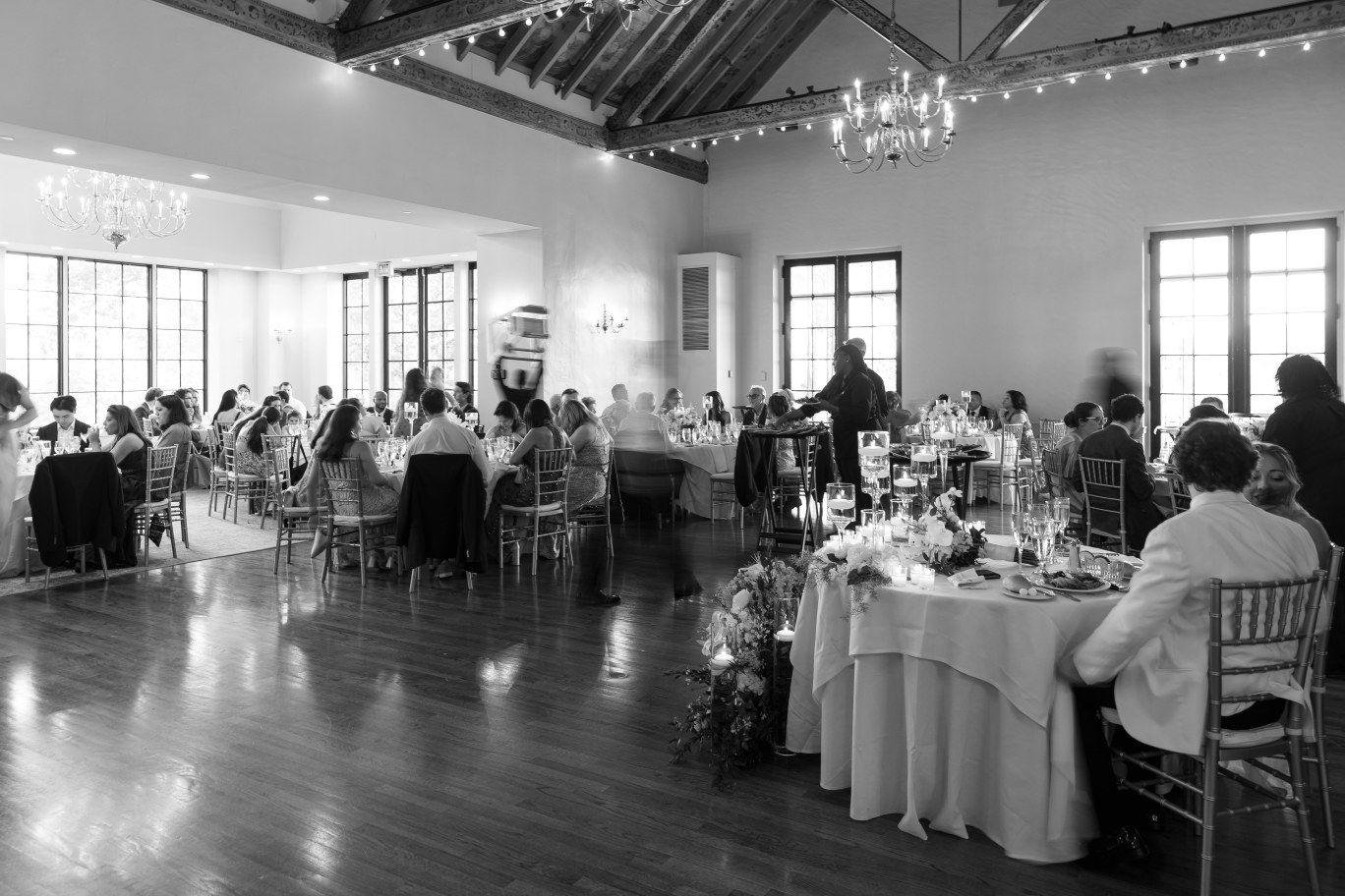 Large indoor event space with multiple banquet tables at a rustic venue features string lights and white linens.