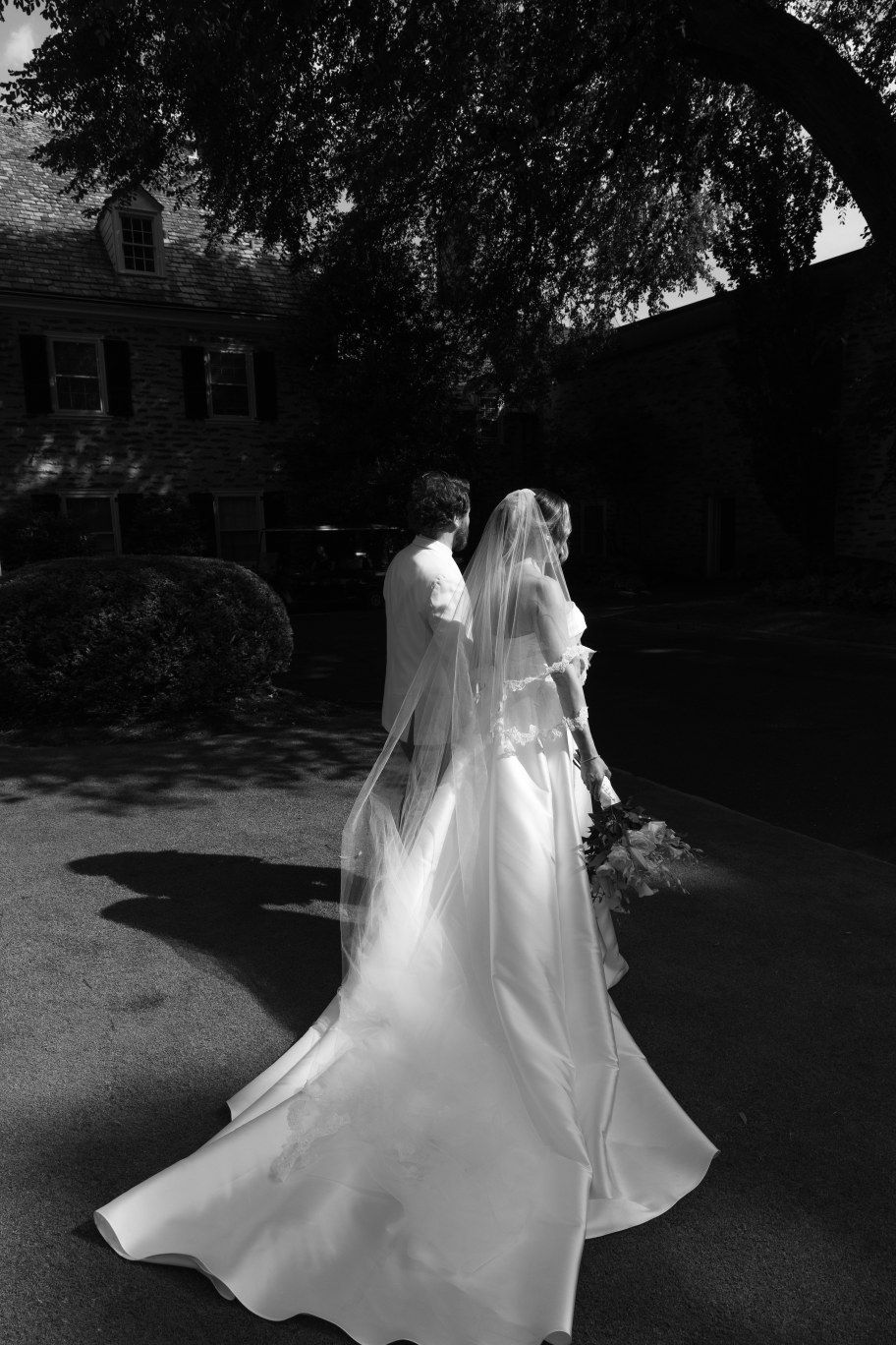 Black and white photograph of a bride in a flowing white wedding gown and veil walking outdoors on a shaded path.
