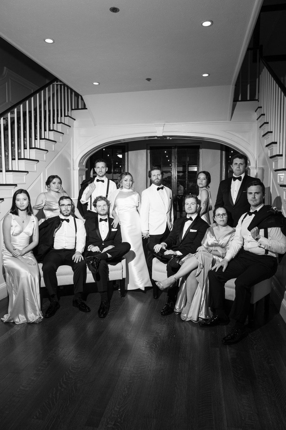 Wedding party members lounge on the floor in formal attire against white walls in an artistic black and white photo.