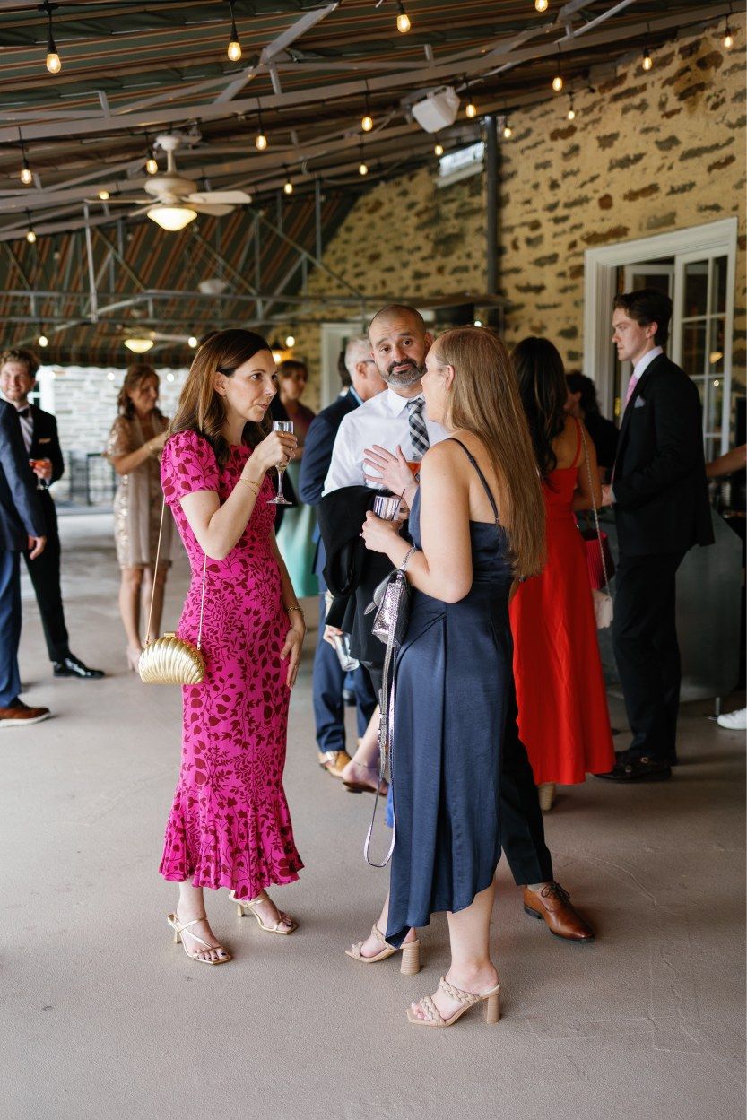 Wedding guests mingle and dance in a rustic stone-walled venue with overhead string lights.