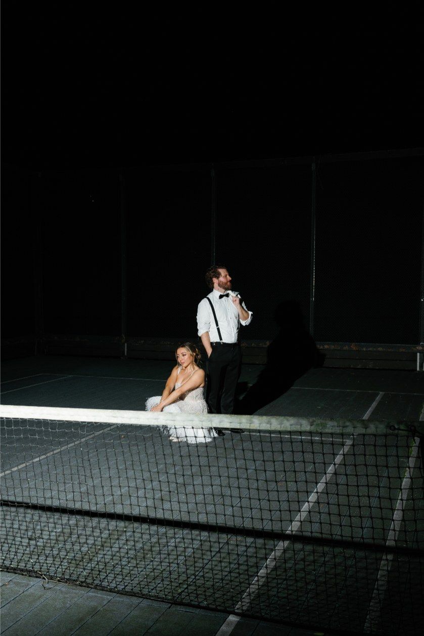 A dramatic wedding portrait taken at night against a tennis court net with striking spotlight illumination.
