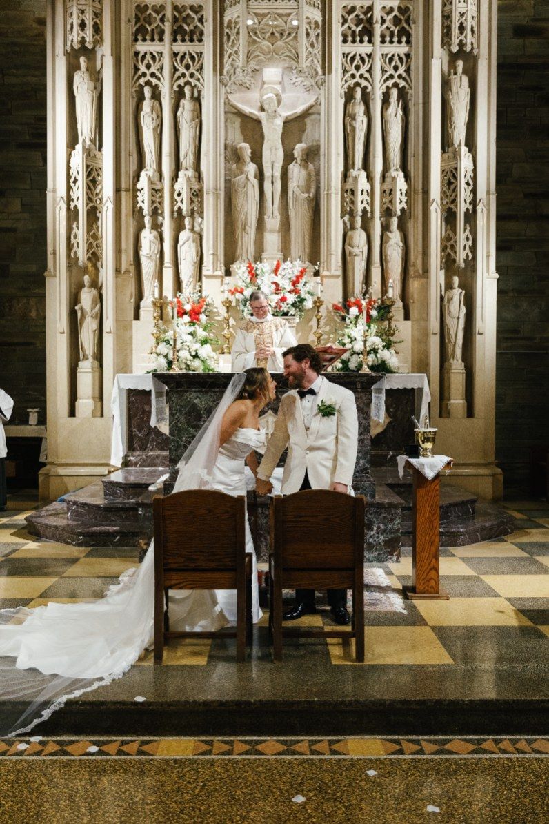 Elegant wedding couple sits together in ornate gothic church with decorative altar and floral arrangements.