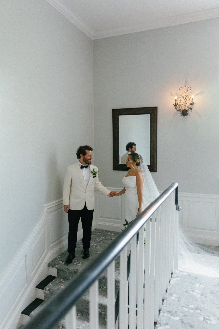 A couple walking down a white hallway with wall sconces during their first look moment at the wedding.