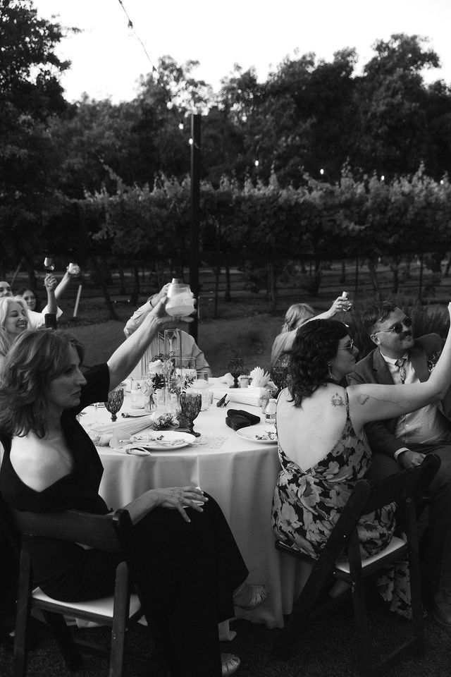Wedding reception guests toast with drinks at outdoor dinner tables at dusk.