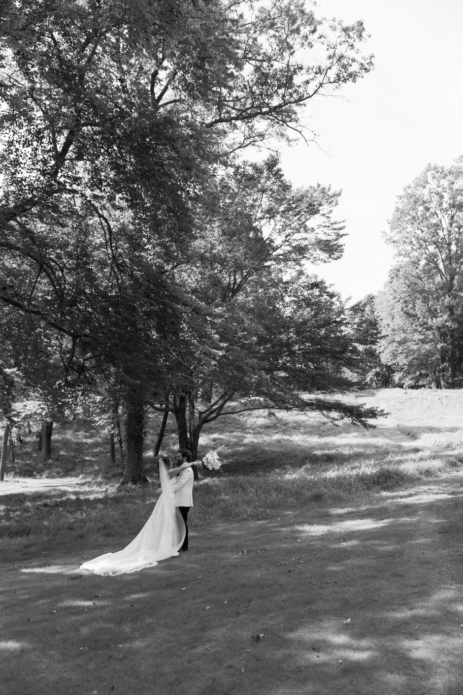 A black and white photo of a couple in wedding attire embracing on a tree-lined hillside path.