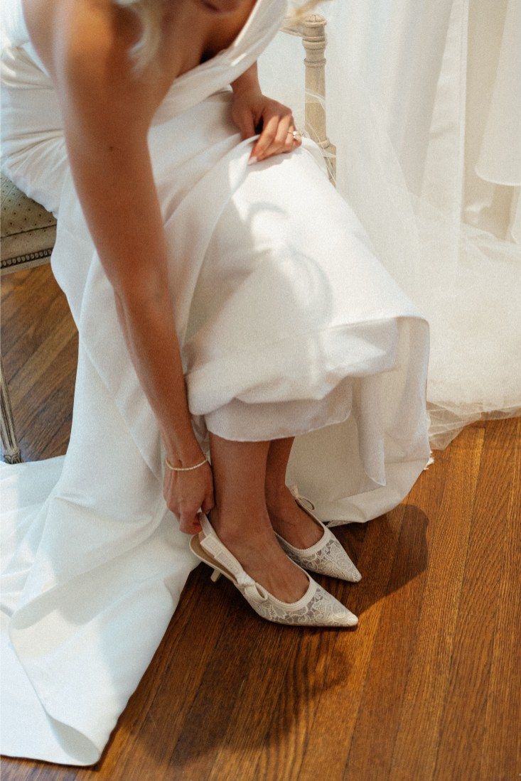 A bride in a white wedding gown adjusts her elegant pointed toe flats while sitting on a wooden floor.