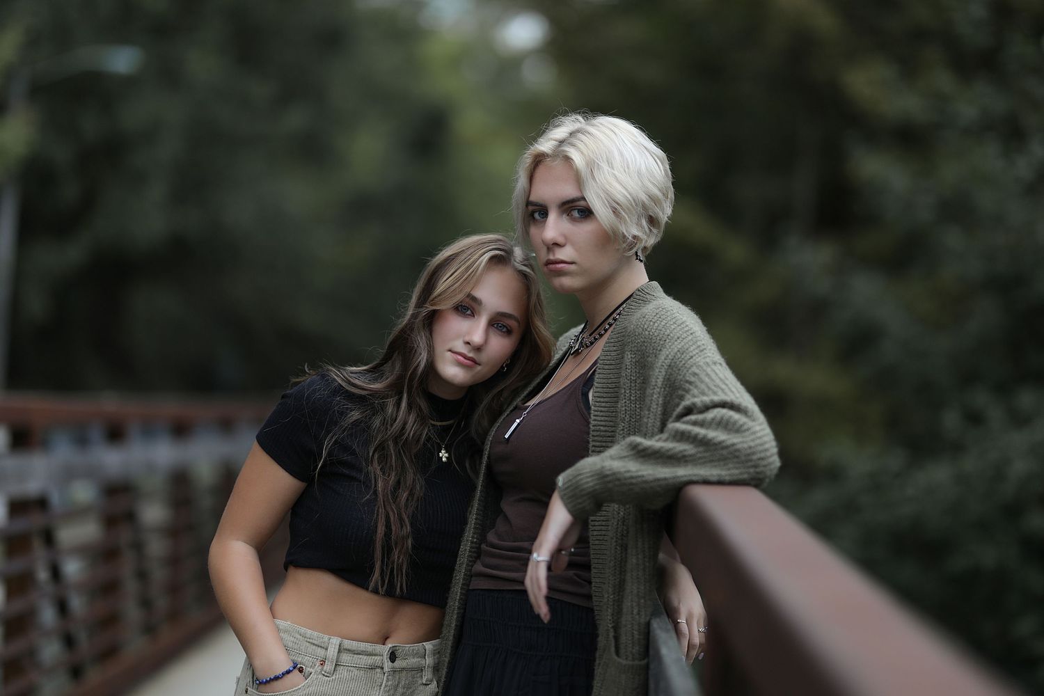 Two people stand together on a wooden bridge railing, wearing casual clothing in an outdoor moody setting.