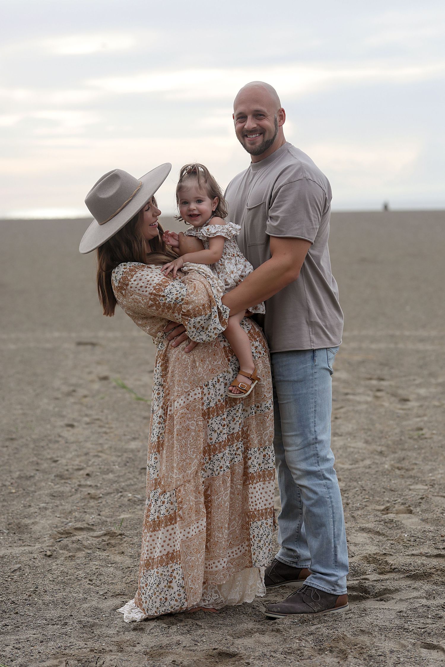 A family of three stands together on an empty beach at sunset wearing casual western-style clothing.