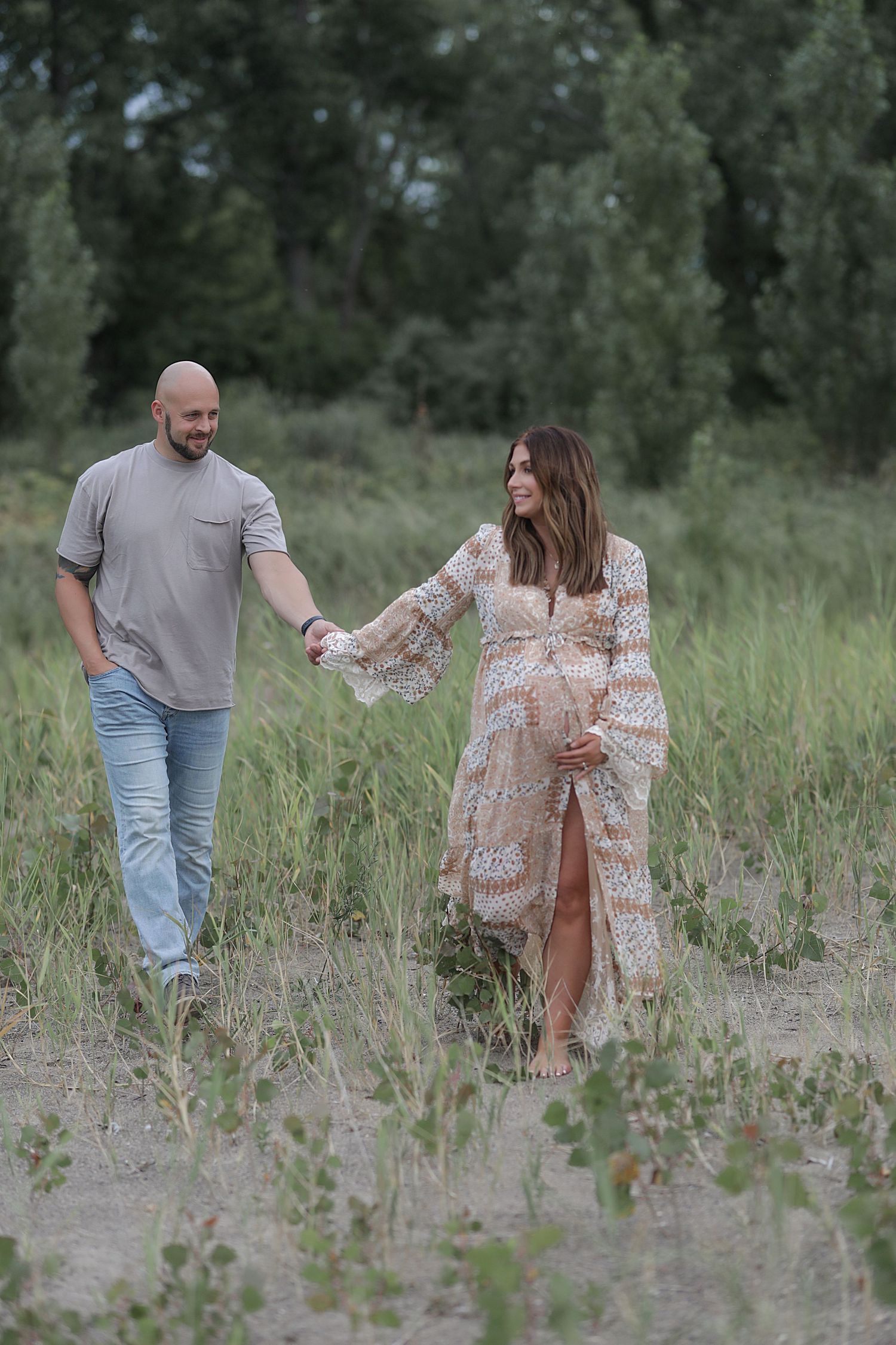 A couple holds hands while walking through tall grass in a romantic maternity photoshoot outdoors.