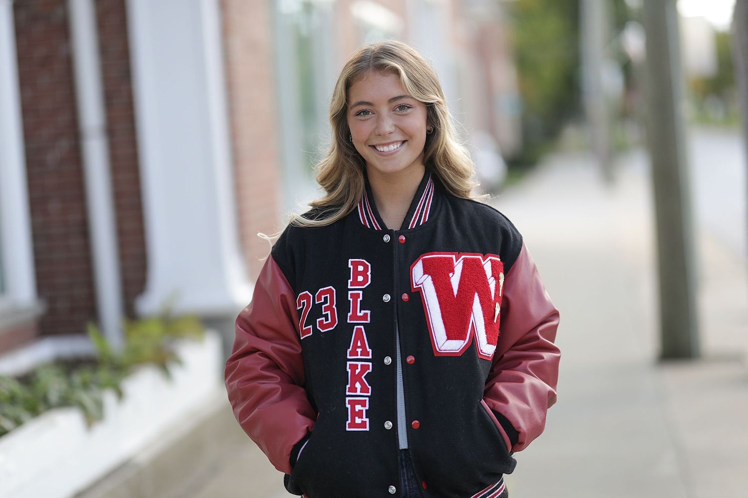 Student wearing a black and red varsity letterman jacket smiles while standing outside on a sidewalk.