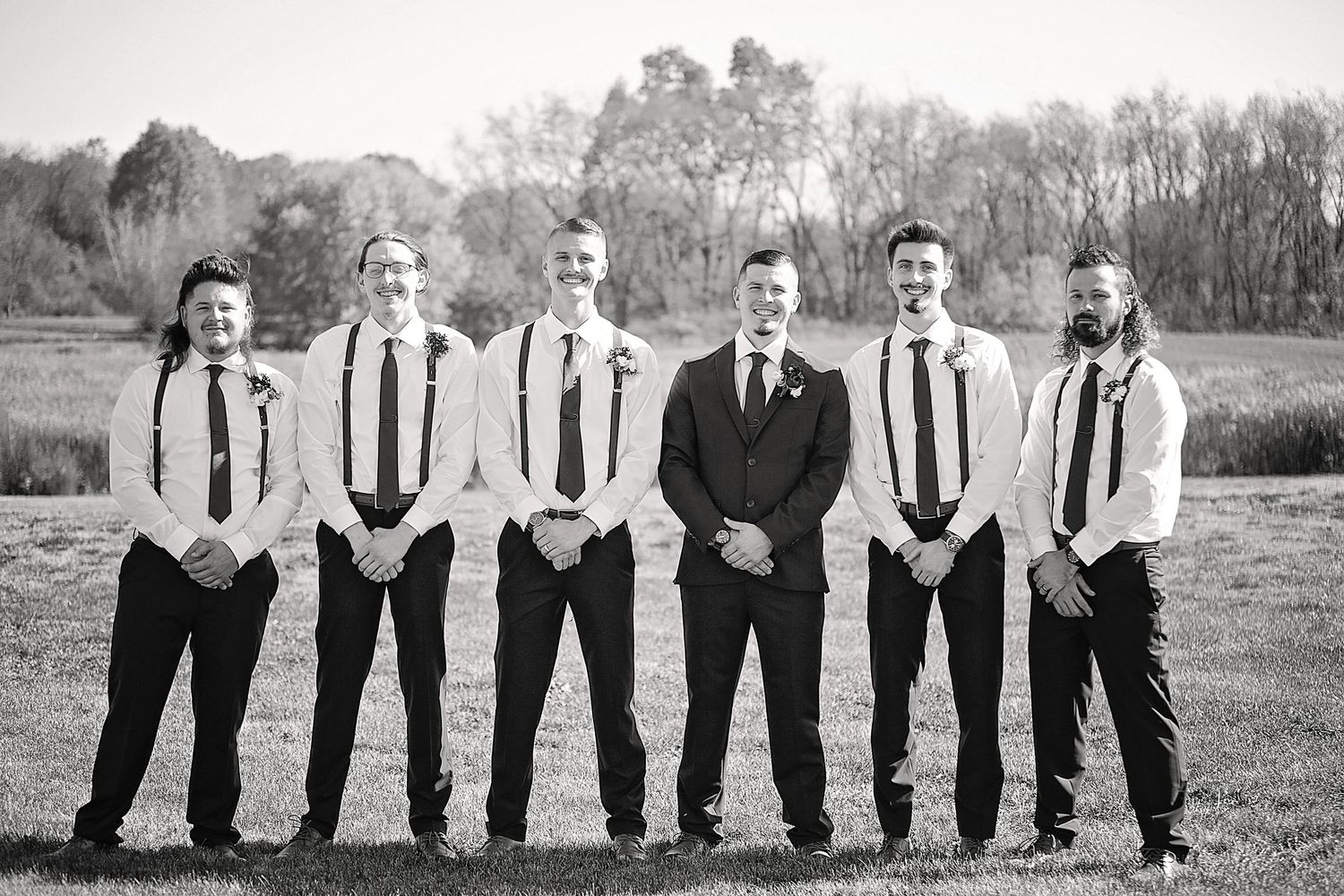 A black and white photo of groomsmen in suspenders and ties posing in a field.
