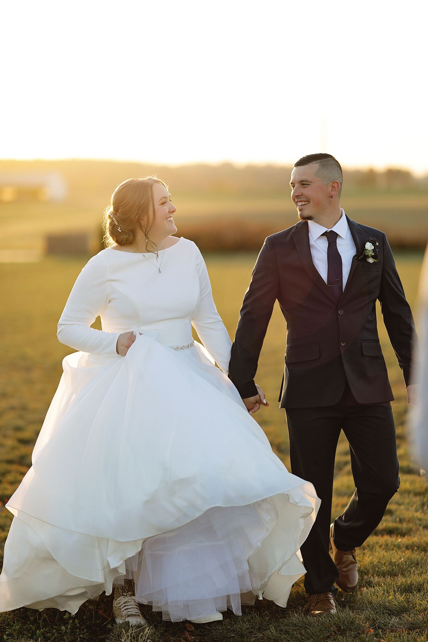 Bride in flowing white dress walking with groom across field at golden hour.
