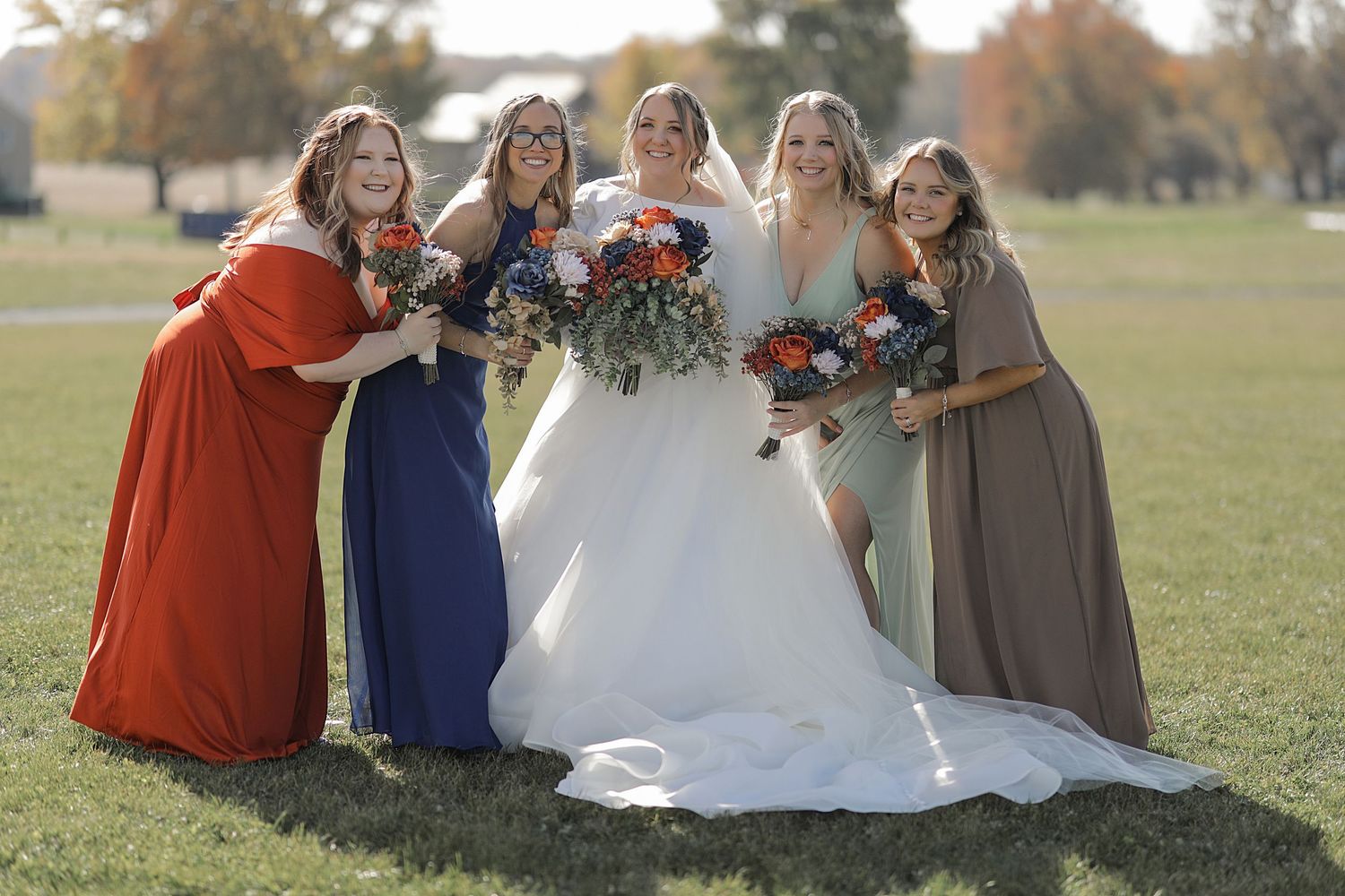 A group poses with colorful bouquets wearing burgundy, blue, and neutral-toned dresses.