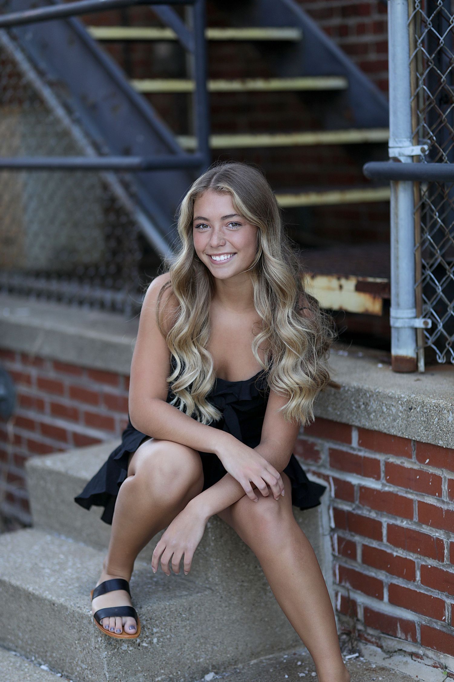 A person in a black dress sits on brick stairs outdoors with long wavy blonde hair and sandals.