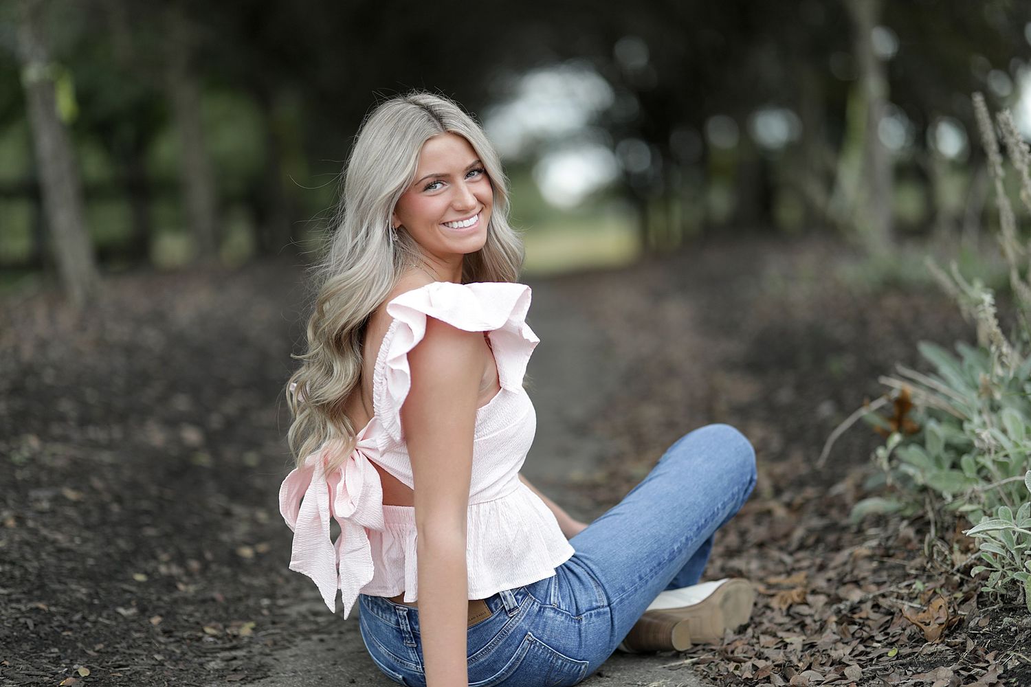 A woman in a white ruffle top and jeans poses casually outdoors in a natural garden setting.