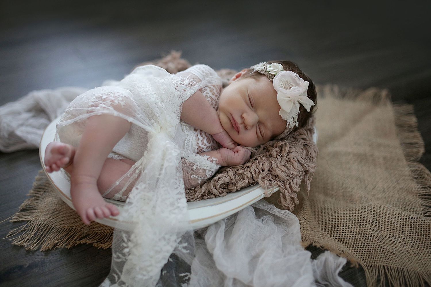 A sleeping baby wearing a white lace dress and headband rests peacefully on textured fabric.
