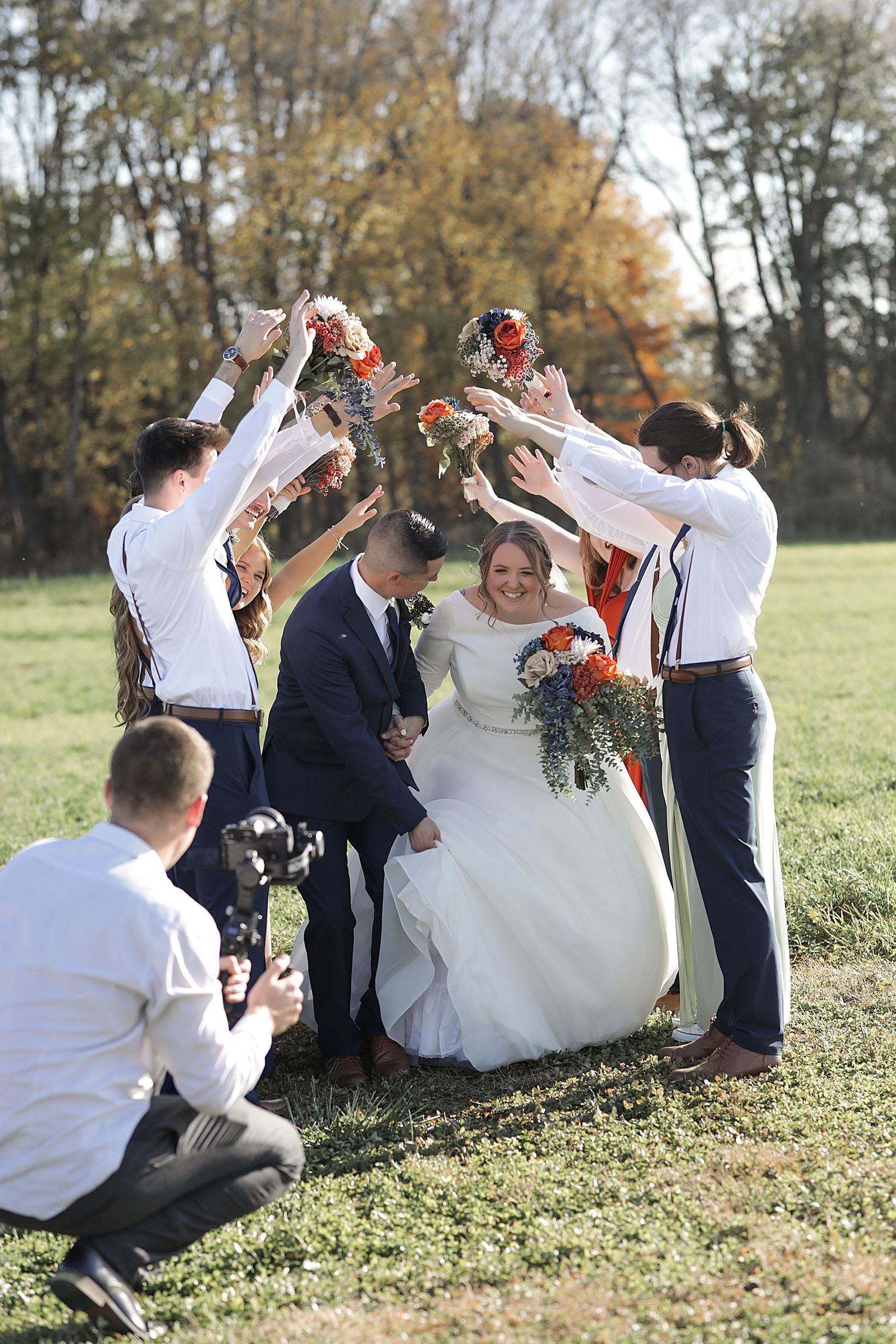 Wedding party members lifting bouquets while photographer captures moment on sunny autumn day.