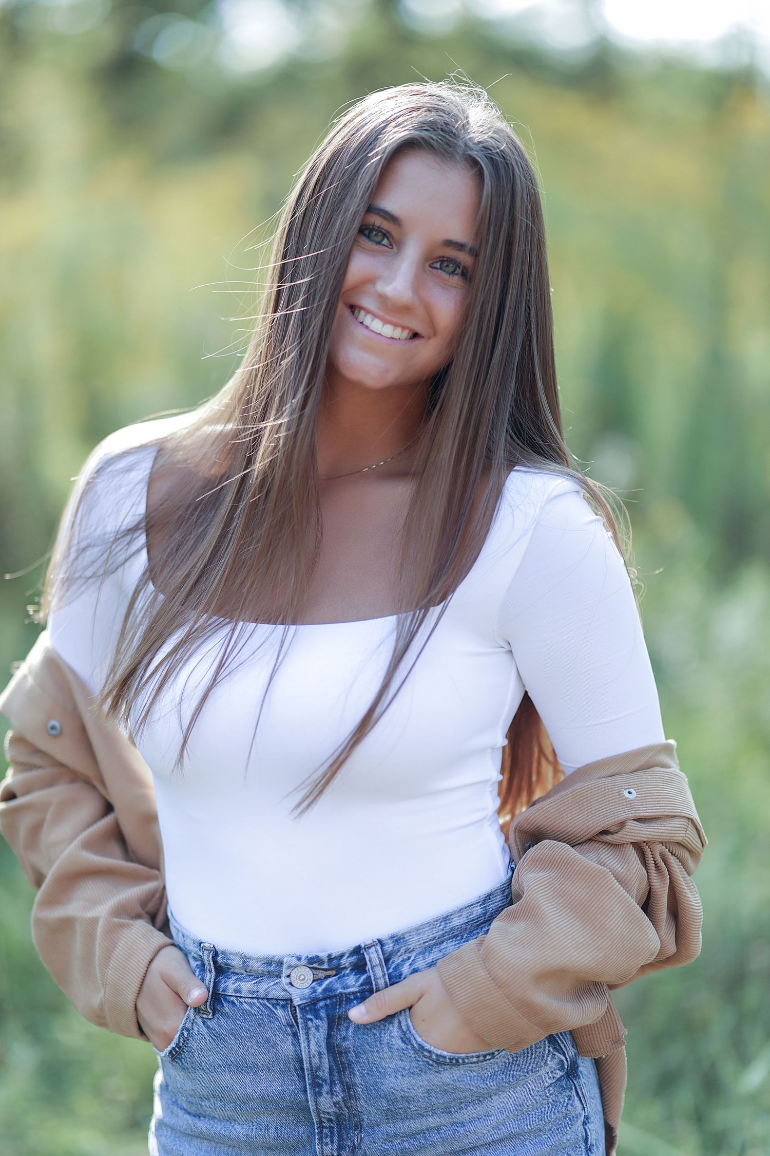 A person with long straight brown hair poses outdoors in a white top, tan jacket and blue jeans with natural lighting.