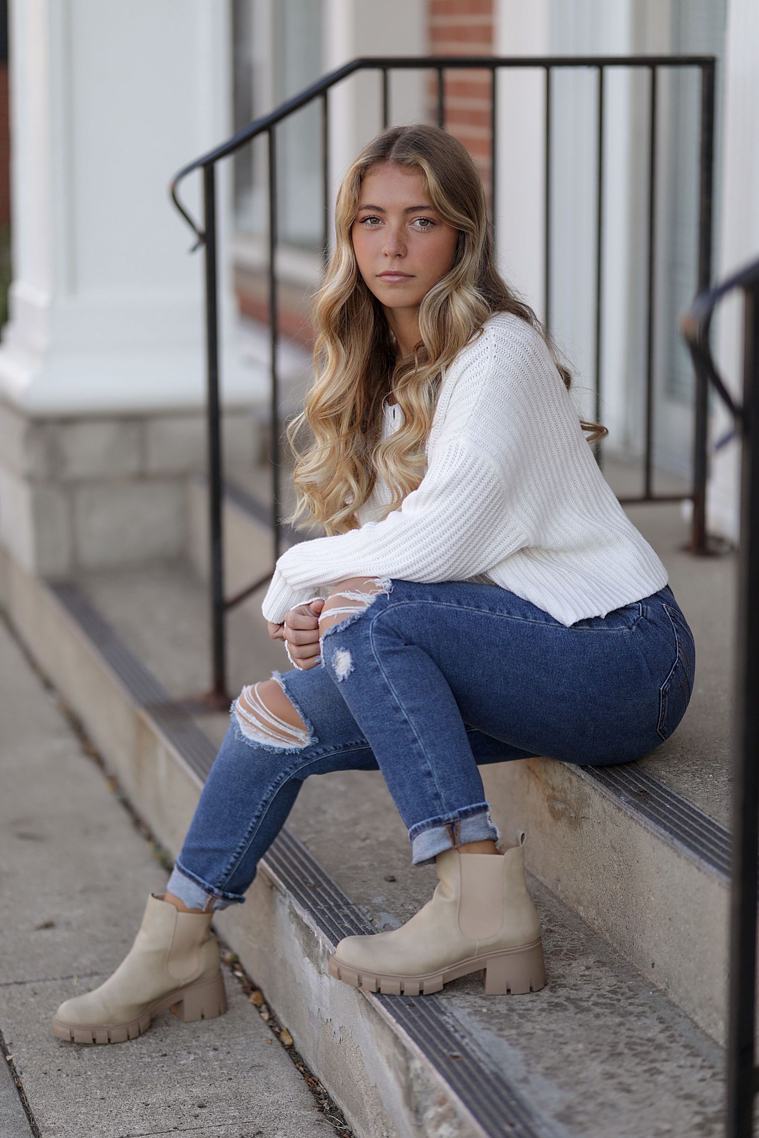 A person in a white sweater and blue jeans sits on concrete steps while wearing beige ankle boots.