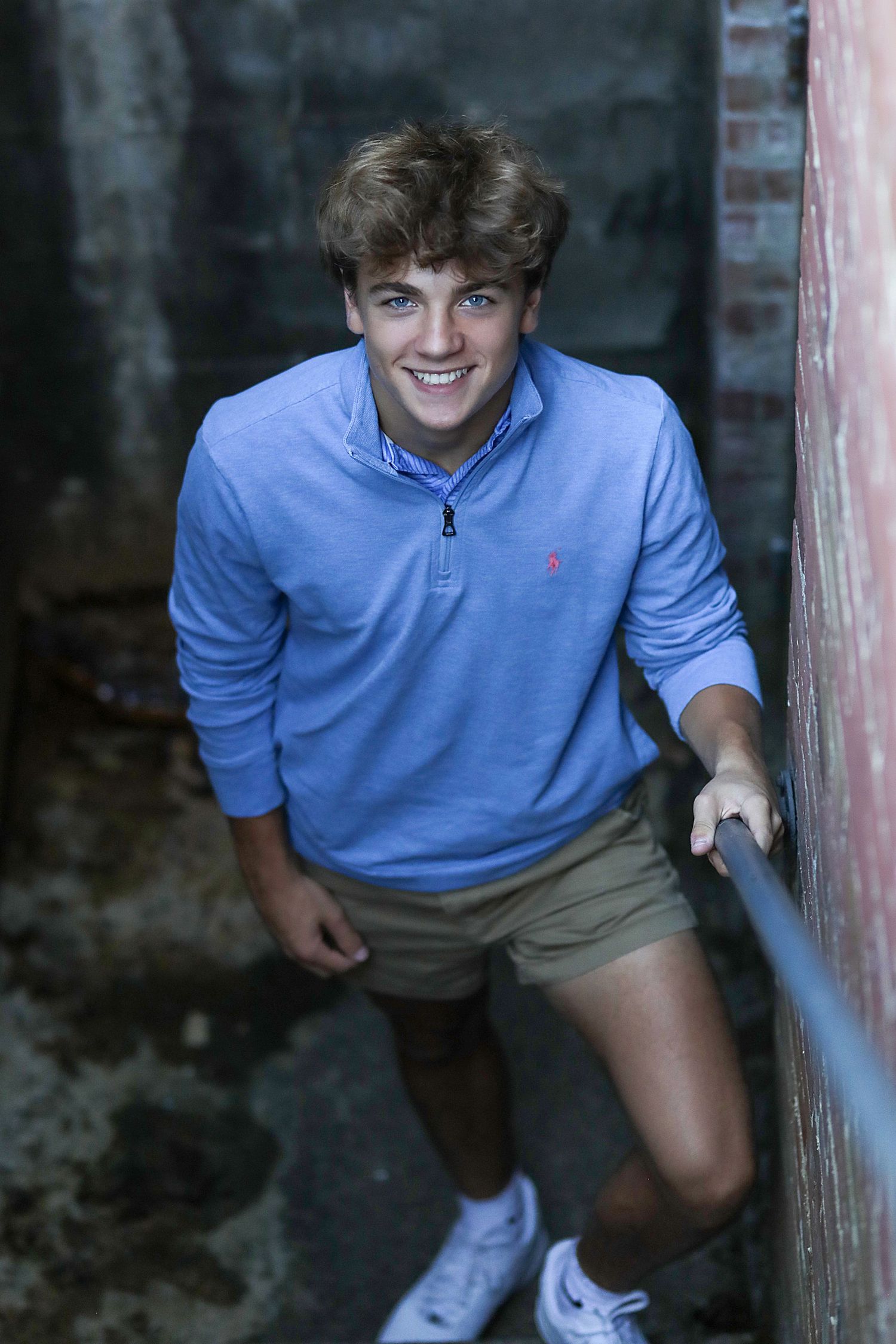 Young person wearing a light blue pullover and khaki shorts poses against a brick wall in casual athletic style.