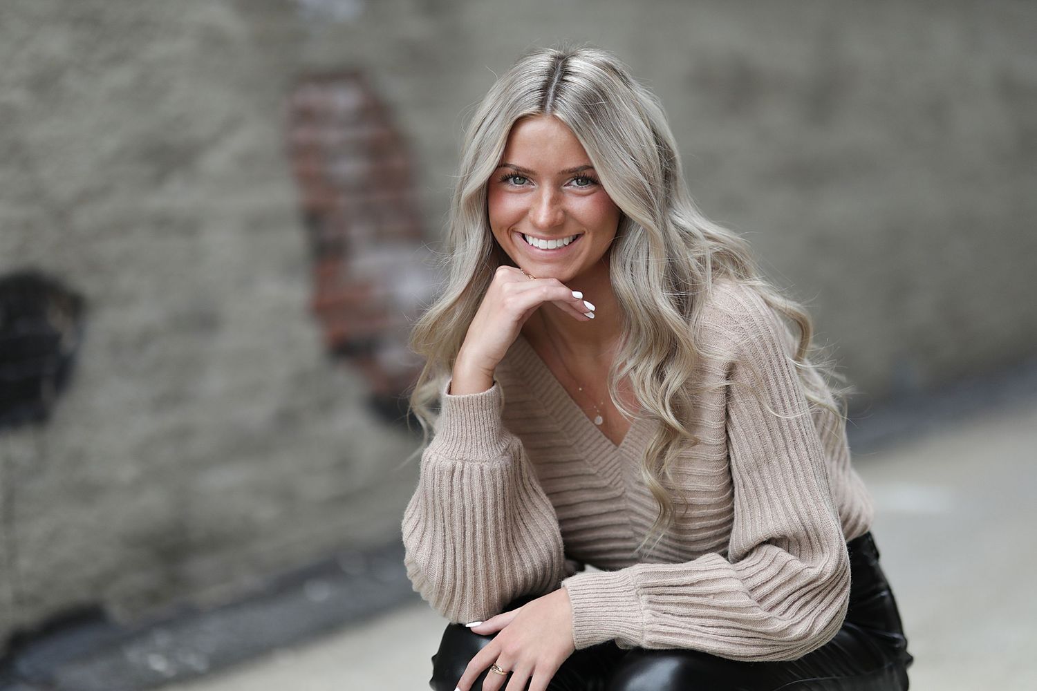 Person in a cozy beige sweater poses and smiles against a stone wall background in a casual outdoor setting.