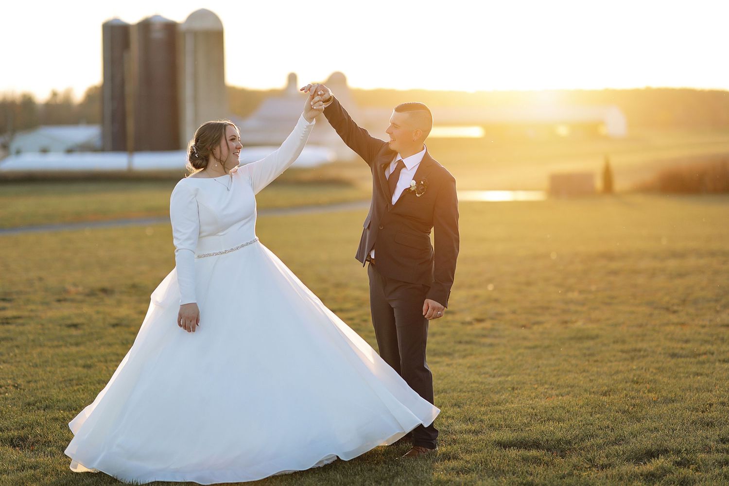 Newlyweds dancing in golden sunset light with rustic farm buildings in background.