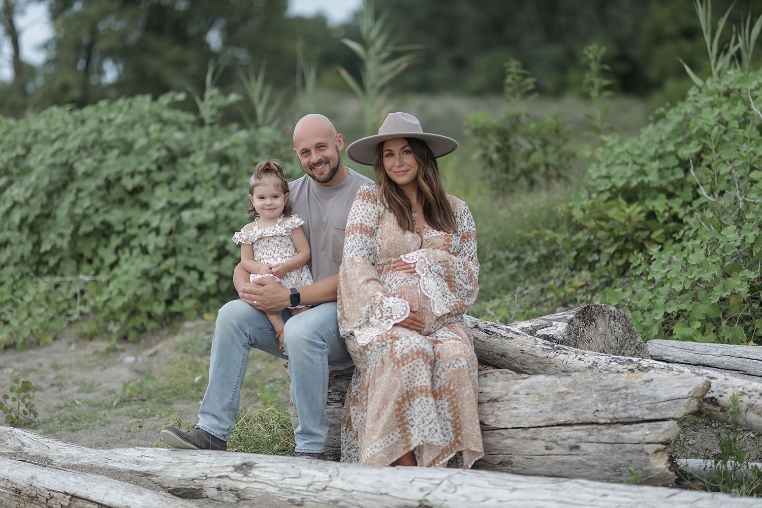 Family wearing neutral colored outfits sitting together on driftwood log in natural outdoor setting during photo session.