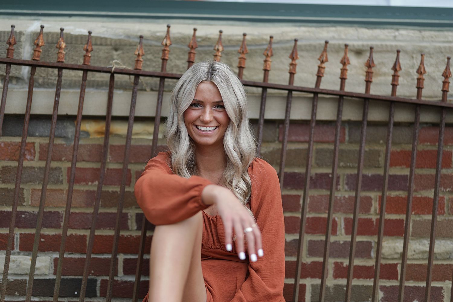 Woman with long silver hair wearing a rust-colored outfit sits smiling by a metal fence at the beach.