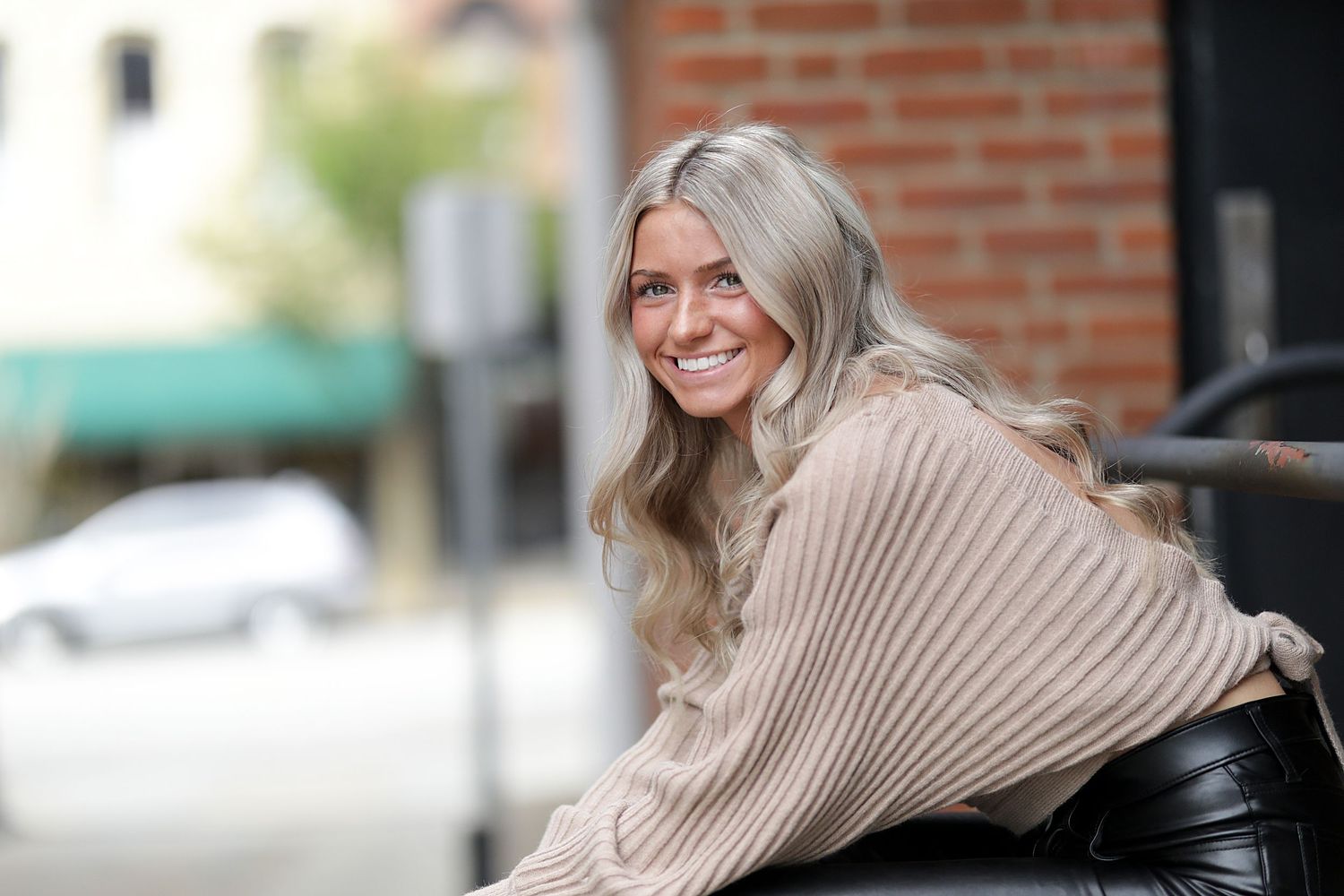 A person in a beige sweater smiles and poses against a brick wall background.