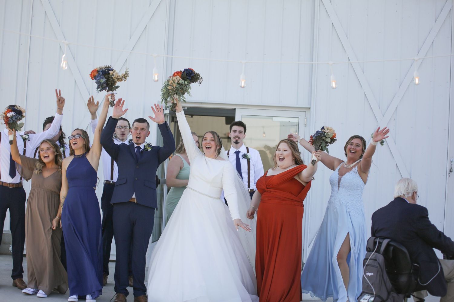 Wedding party celebrating with raised arms against white barn backdrop.