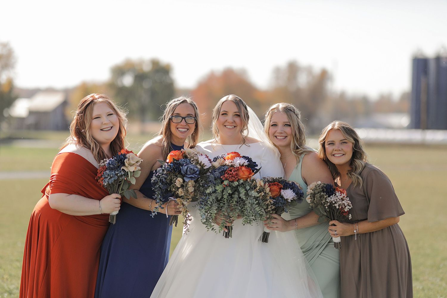 Wedding party in colorful dresses holding floral bouquets posing on grassy field.