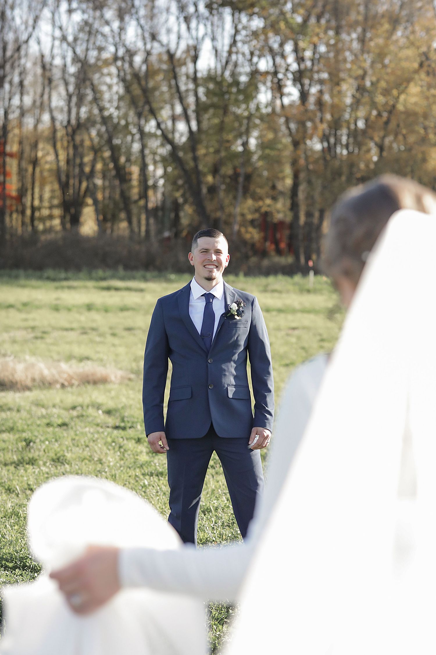 Groom in navy suit stands in grassy field during first look wedding photos with flowing white veil in foreground.