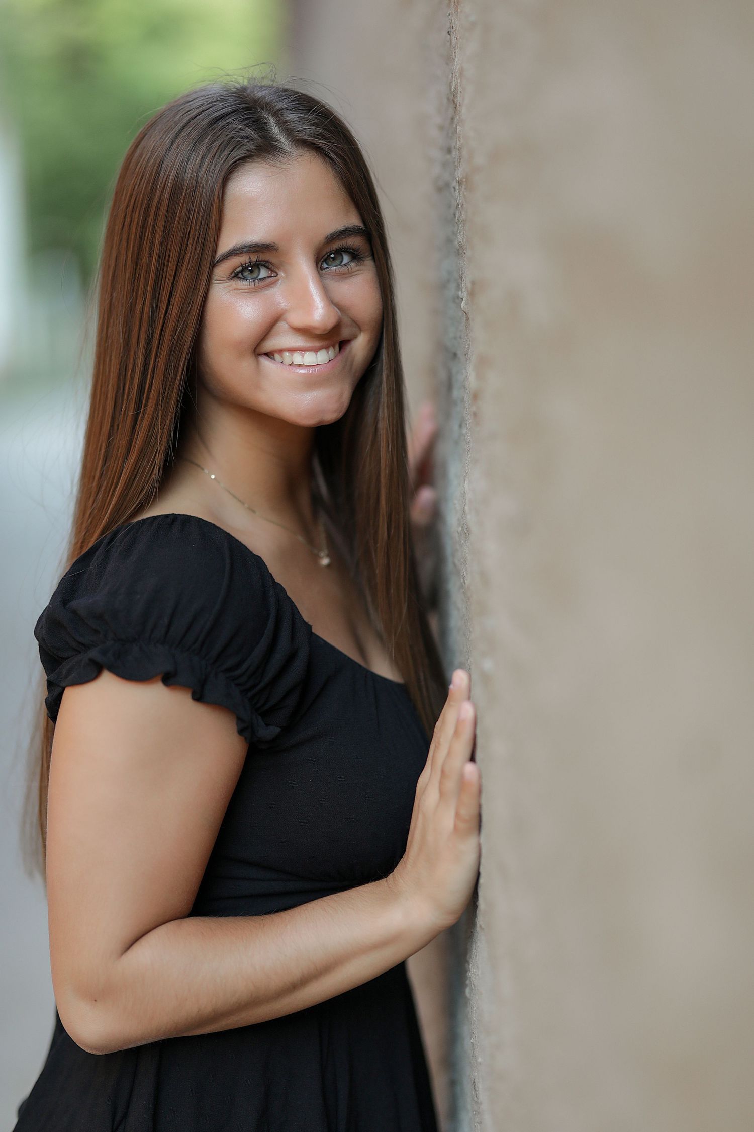 Portrait of someone in a black dress leaning against a wall outdoors with natural lighting and a warm smile.