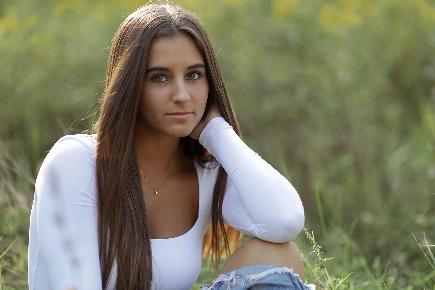 Young woman in white sweater sitting outdoors in a natural setting with long brown hair.