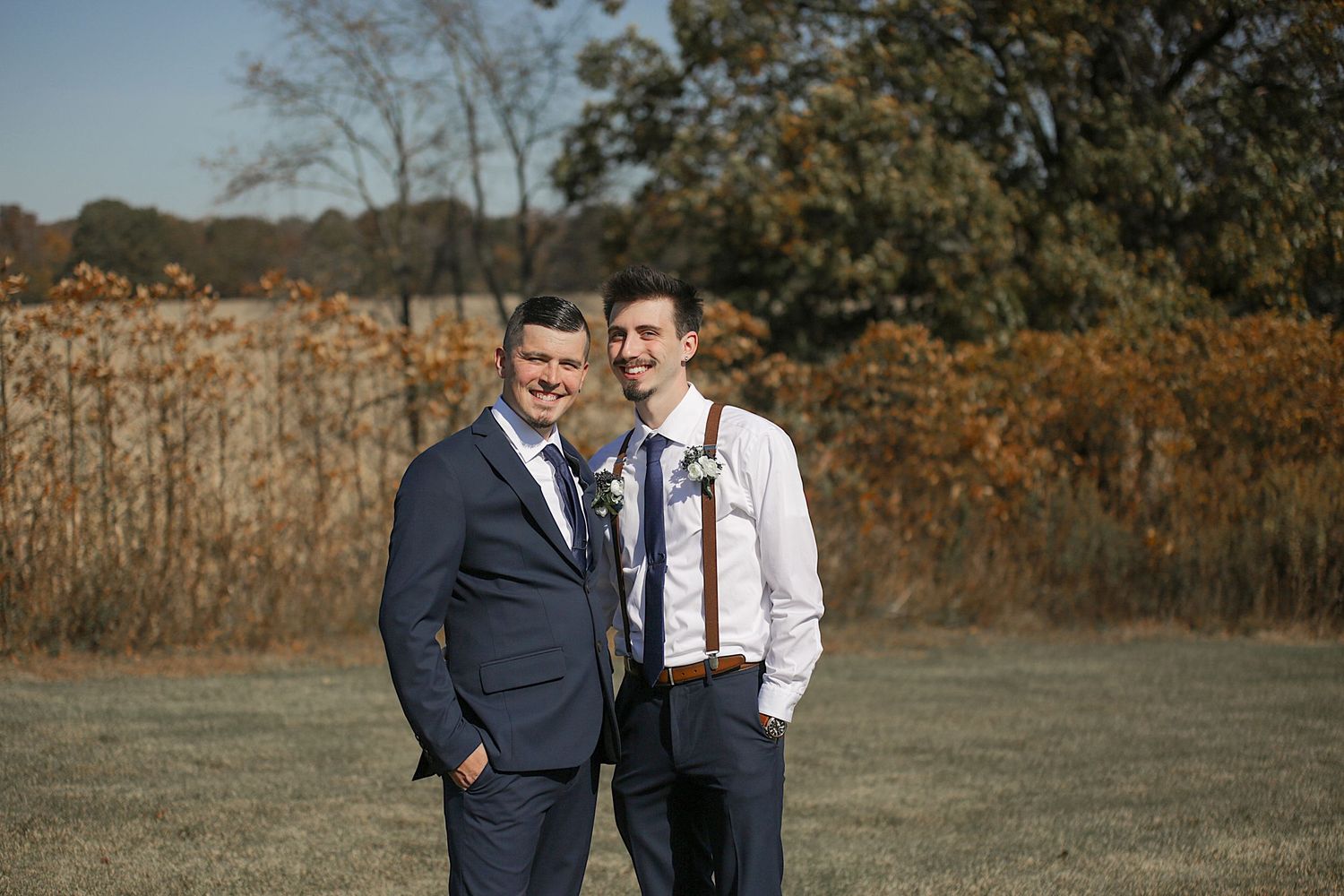 Two wedding guests share a lighthearted moment in a fall field with golden foliage.