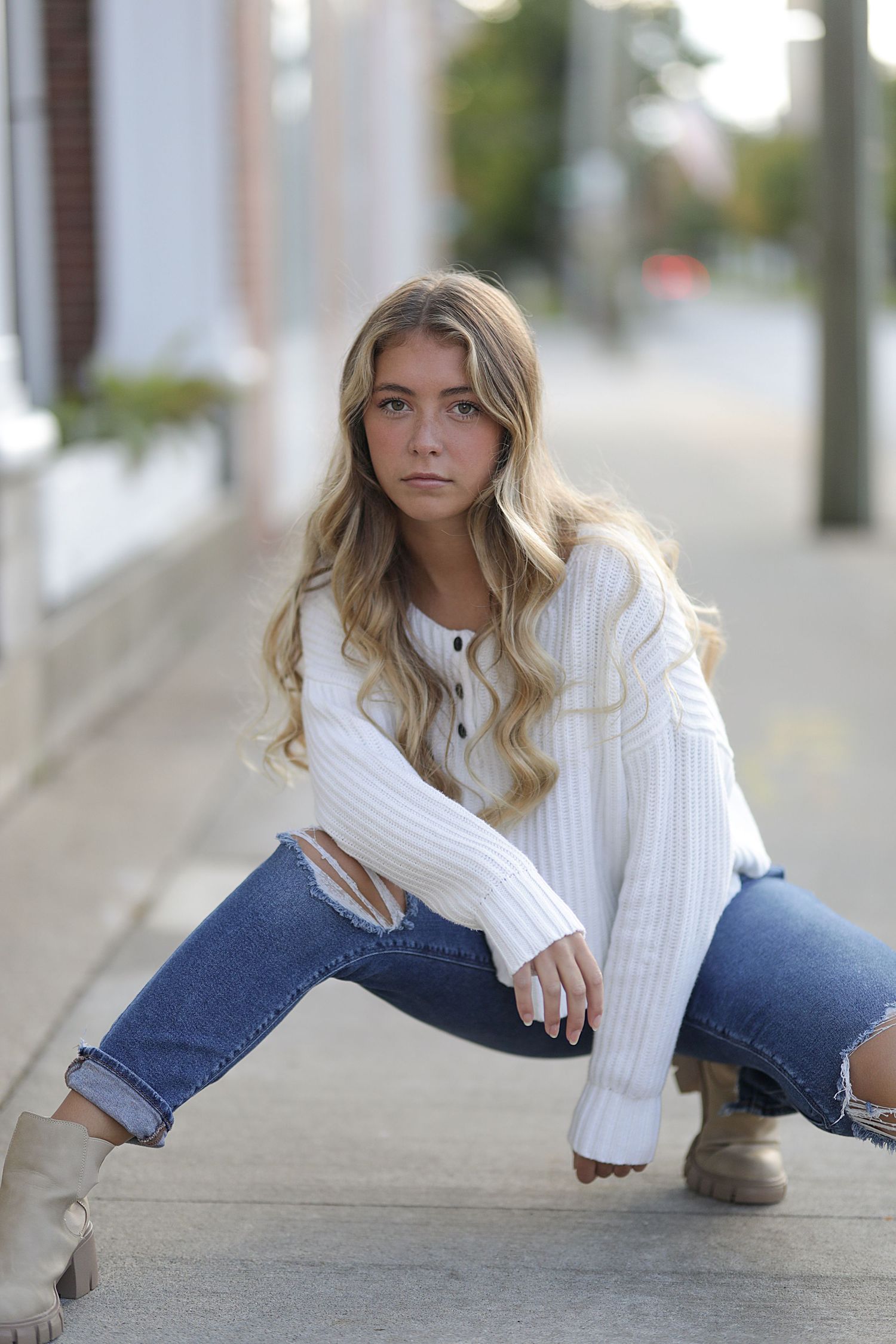 Fashion model poses on sidewalk wearing white knit sweater and distressed jeans in urban setting.