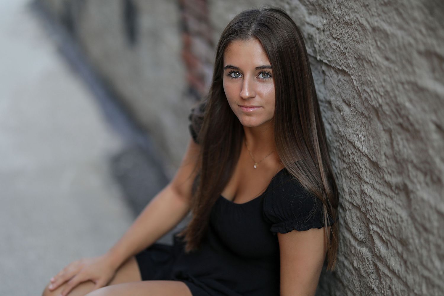 Portrait of woman with long dark hair wearing a black top while leaning against textured wall outdoors.