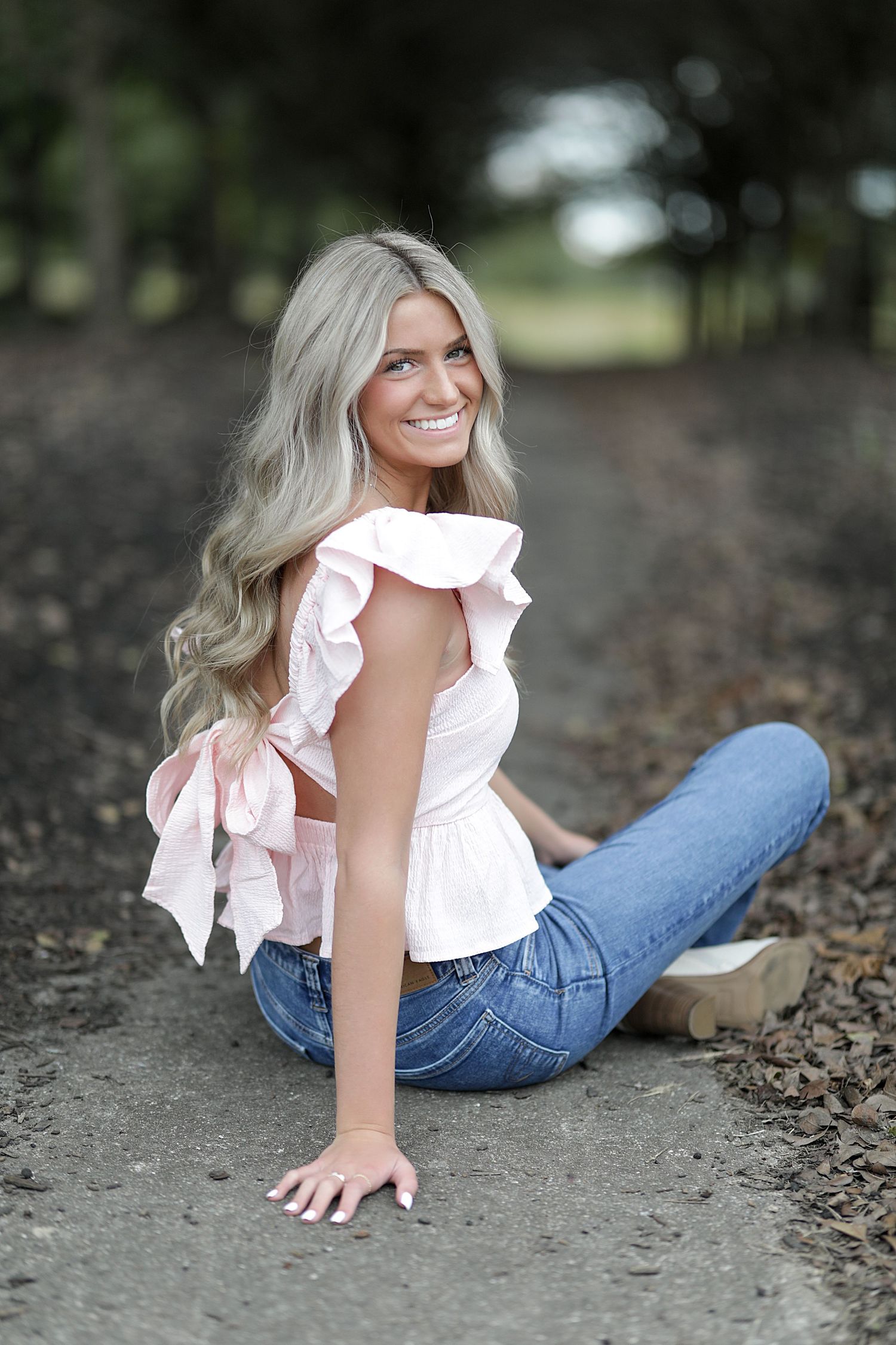 A relaxed outdoor pose in a white ruffled top and blue jeans sitting on a rustic ground with trees in background.