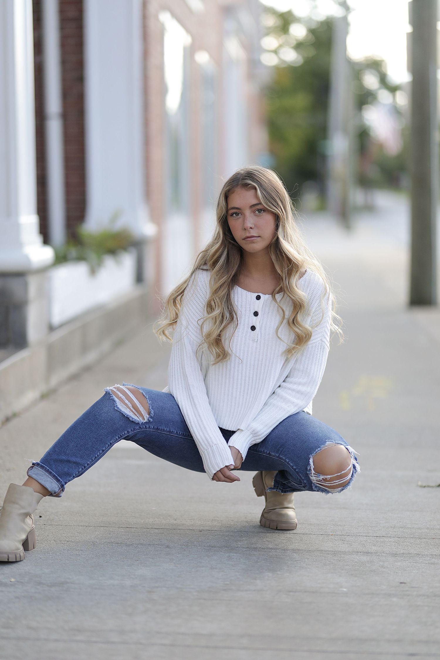 Model wearing white knit sweater and distressed jeans crouches on sidewalk in stylish casual autumn fashion pose.