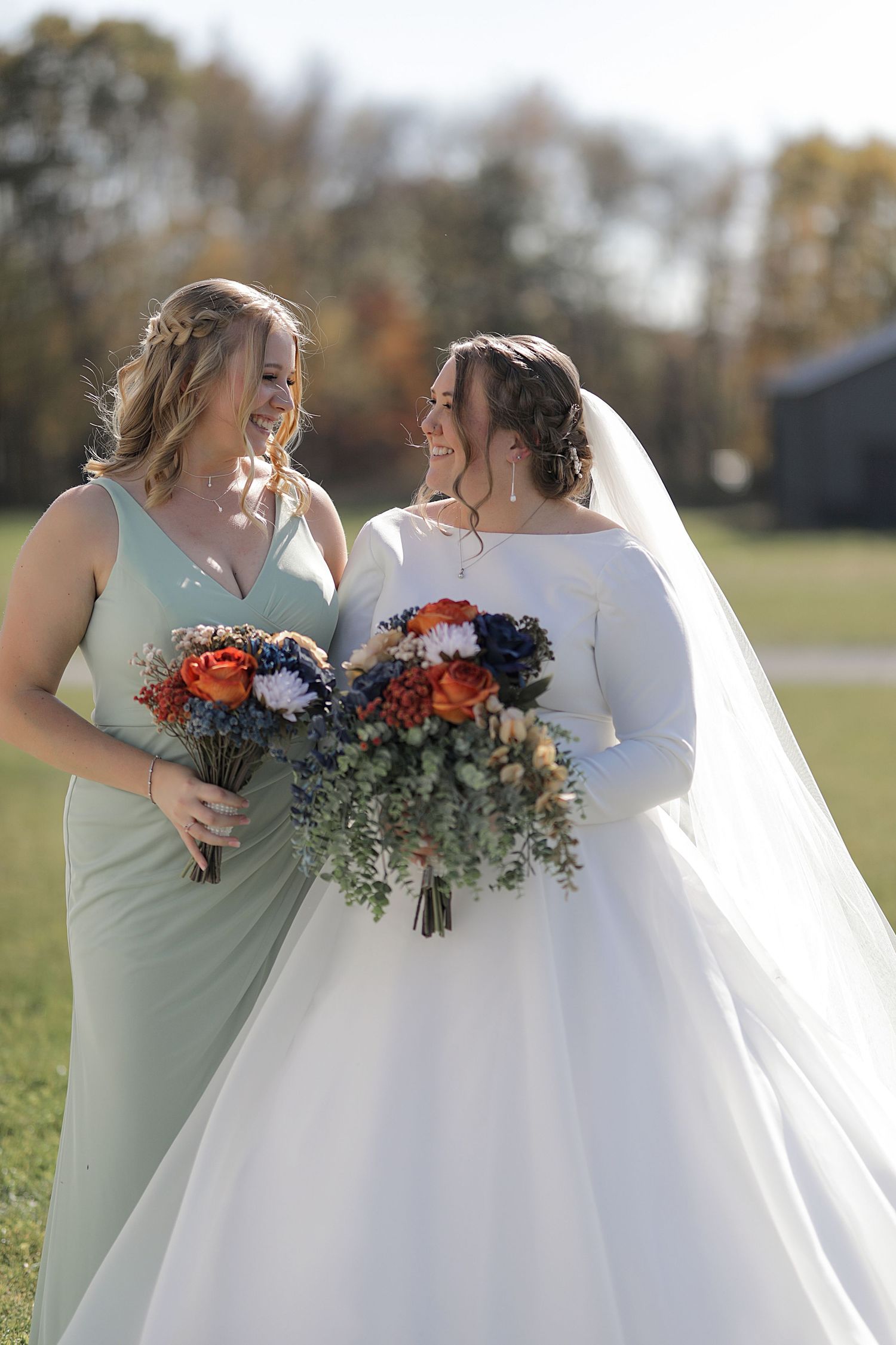 Wedding party members in sage green and white dresses share joyful moments while holding vibrant orange and blue bouquets outdoors.