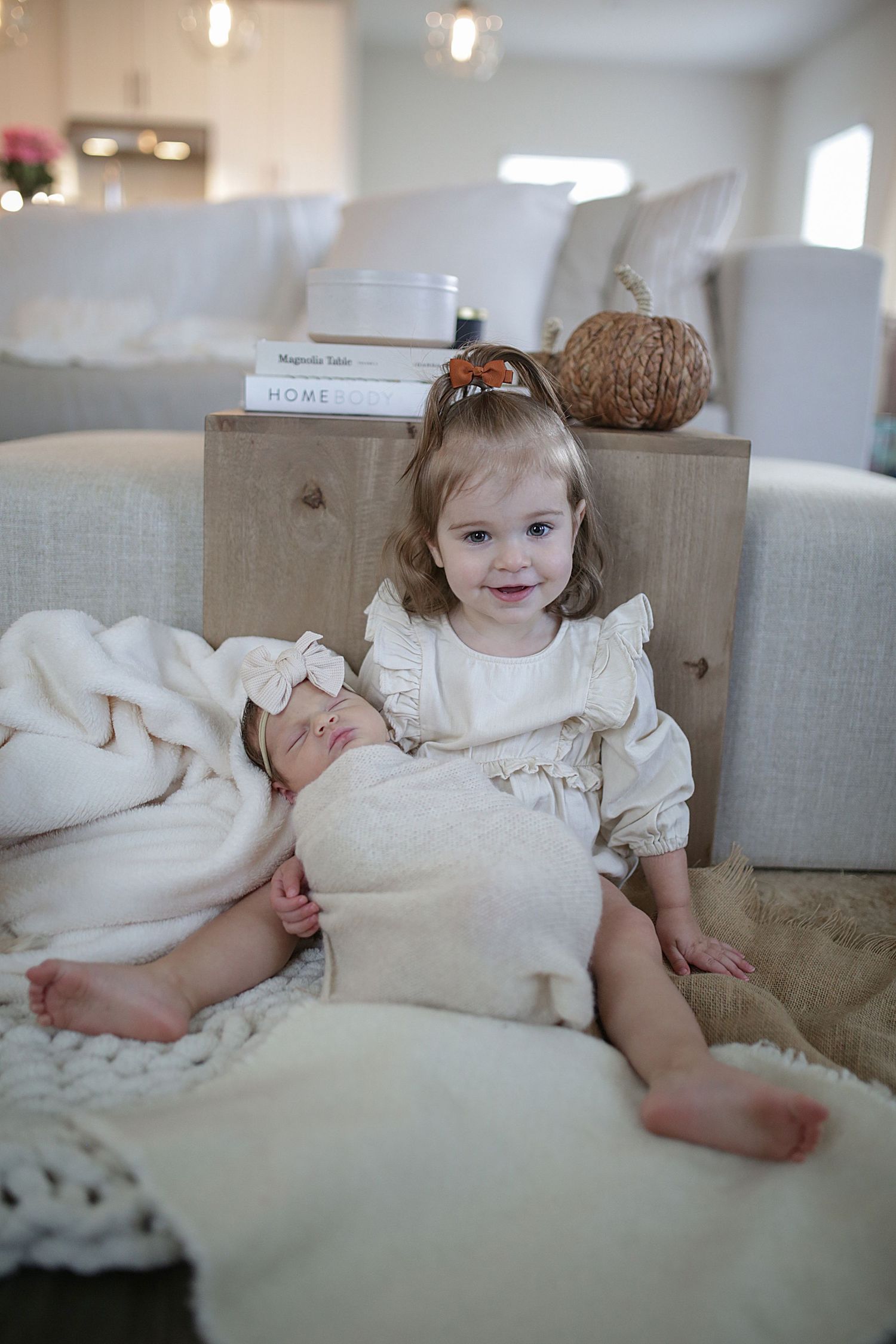 A small child relaxes on a plush cream-colored couch wearing a white ruffled dress in a bright interior space.