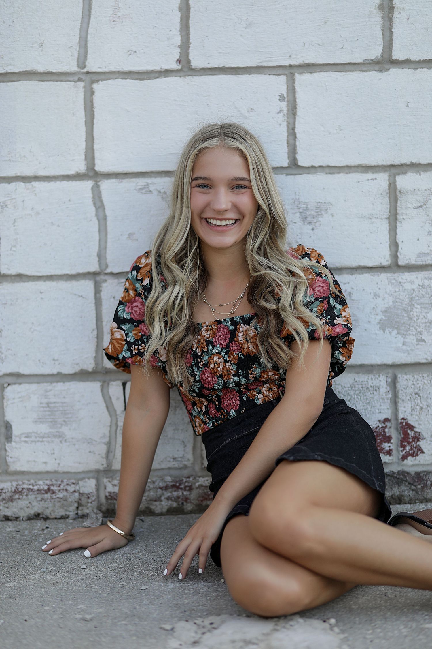 Young woman in floral top and black shorts sits smiling against white brick wall in casual outdoor portrait.