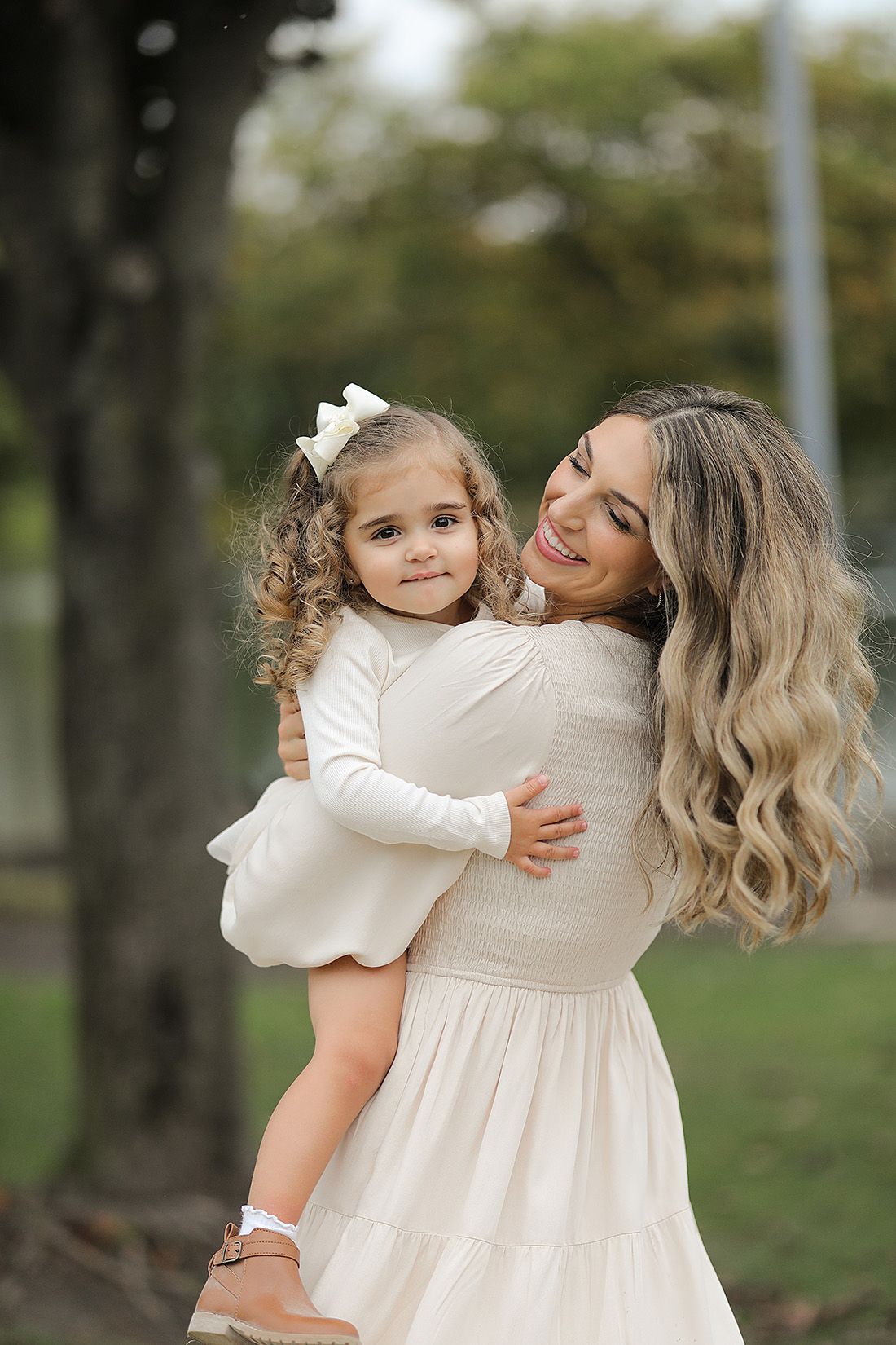 Mother and daughter in matching cream dresses share loving embrace during outdoor portrait session.